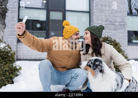 joyeux homme en bonnet tricoté prenant selfie avec une petite amie gaie et chien berger australien en hiver Banque D'Images