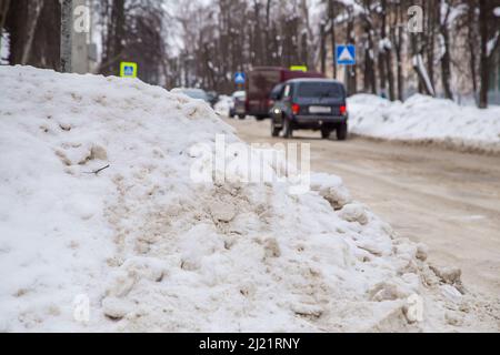 Une profonde dérive de neige au bord de la route sur fond de rue de la ville. Sur la route se trouve de la neige sale dans des tas hauts. Paysage urbain d'hiver. Jour d'hiver nuageux, lumière douce. Banque D'Images