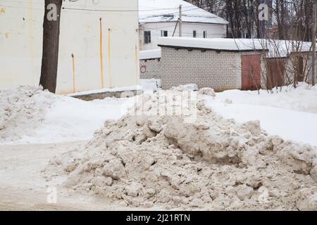 Une grande dérive de neige au bord de la route sur fond de rue de la ville. Sur la route se trouve de la neige sale dans des tas hauts. Paysage urbain d'hiver. Jour d'hiver nuageux, lumière douce. Banque D'Images