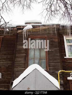 Des glaçons transparents d'hiver sont suspendus sur le bord du toit. Sur le fond du mur en bois de la vieille maison. Grandes cascades, même de belles rangées. Jour d'hiver nuageux, lumière douce. Banque D'Images