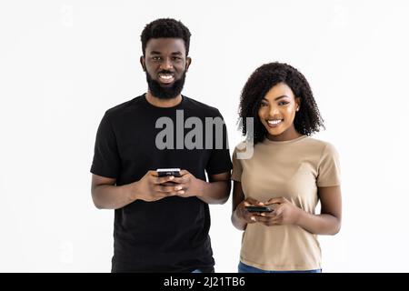 Un couple africain séduisant tient des smartphones qui s'amusent sur Internet, isolés sur fond gris de studio Banque D'Images