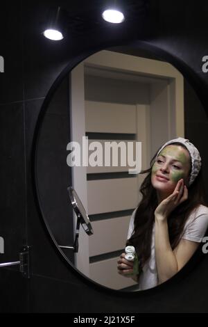 Femme en bandeau appliquant un masque en argile vert sur son visage dans une salle de bains intérieure sombre à la maison devant un miroir. Soin naturel de la peau. Fille prenant soin d'oi Banque D'Images