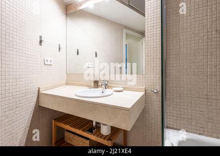 Salle de bains avec lavabo en porcelaine blanche sur comptoir en marbre crème et mosaïque, miroir sans cadre fixé au mur et armoire en bois sous le comptoir Banque D'Images
