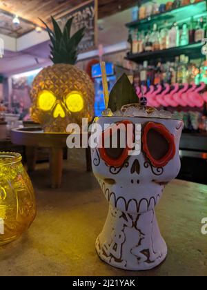 Un cocktail tiki dans une tasse en céramique de crâne de sucre au-dessus d'un bar. Derrière la boisson se trouve une lumière de crâne d'ananas. Banque D'Images