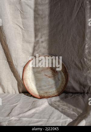 Plaque en céramique faite à la main photographiée sur du linge biologique, partiellement cachée dans l'ombre et la lumière Banque D'Images