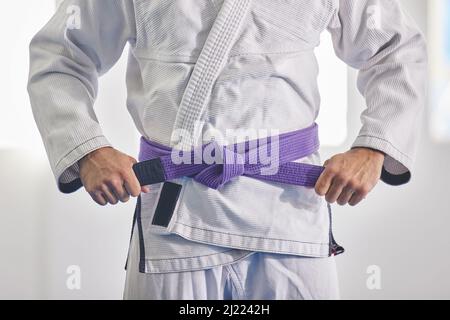 Par une ceinture violette. Coupe courte d'un homme méconnaissable nouant une ceinture mauve autour de la taille du poing tout en jiu jitsu gi complet. Banque D'Images