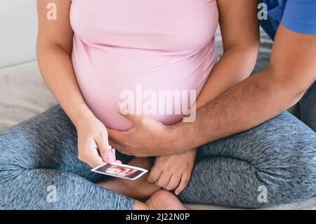 l'homme en gros plan touche le ventre et la femme enceinte tient l'échographie assis sur le lit, heureux couple caucasien regardant l'échographie imprimée de leur bébé. Banque D'Images
