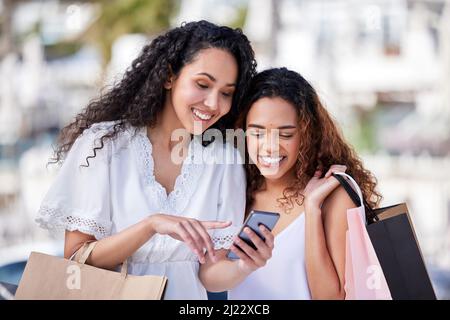 Les meilleurs amis partagent les meilleures offres avec vous. Prise de vue de deux jeunes femmes utilisant un smartphone tout en faisant des achats sur un fond urbain. Banque D'Images
