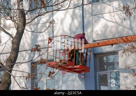 Le constructeur d'un panier de grue répare le revêtement d'une structure métallique sur le mur de la façade d'un bâtiment. Banque D'Images