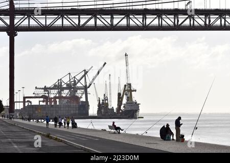 Les gens sur la promenade et les pêcheurs à côté de la rivière Tage près du pont 25 de Abril avec port industriel en arrière-plan, Lisbonne Portugal. Banque D'Images