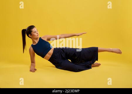 Vue avant de la jeune fille flexible couchée sur le côté, levant le bras et la jambe, en regardant vers l'avant. Brunette femme portant un costume de sport bleu étirant, pratiquant la pose de yoga. Concept de yoga faire. Banque D'Images