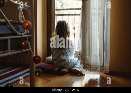 Enfant assis sur le sol et jouant à la maison. Ambiance rêveuse et ensoleillée. Fille regardant par la fenêtre de l'arrière. Un adorable enfant qui attend pour une promenade Banque D'Images