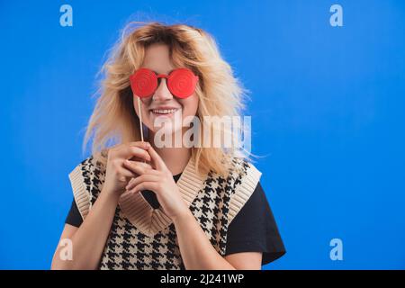 Une jeune femme européenne espiègle qui se grince et qui la couvre les yeux avec de faux verres rouges sur fond bleu. Prise de vue en studio isolée. Concept de fête. Photo de haute qualité Banque D'Images