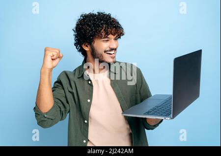Joyeux garçon arabe ou indien dans des vêtements décontractés, étudiant ou indépendant, tient un ordinateur portable ouvert dans sa main, se réjouit de gagner, gesturant avec son poing, debout sur un fond bleu isolé Banque D'Images
