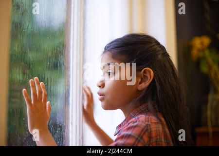 J'espère qu'il cesse de pleuvoir pour que je puisse aller dehors et jouer. Photo d'une petite fille mignonne regardant par la fenêtre un jour de pluie. Banque D'Images