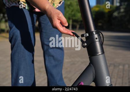 Femme méconnue qui déplie son scooter électrique ; photo détaillée de sa main qui règle le levier du mât. Concepts d'énergie électrique, mobi vert Banque D'Images