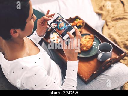 Faites-le pour le gramme. Prise de vue rognée d'une jeune femme attirante utilisant son téléphone portable pour prendre une photo de son plateau de petit déjeuner à la maison. Banque D'Images