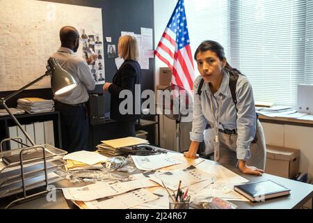 Jeune détective hispanique sérieuse, femme, se pencher sur le lieu de travail avec des documents sur l'arrière-plan de deux collègues communiquant Banque D'Images