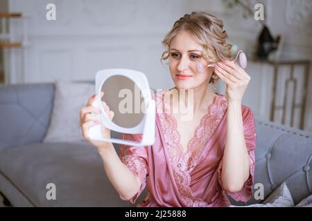 Femme utilisant le rouleau facial. Sculpture, soulèvement et soins anti-anxiété. Soins de la peau et beauté à la maison. Femme utilisant un masseur à rouleaux en quartz apaisant tout en étant assise sur un canapé dans le salon, regardant le miroir Banque D'Images