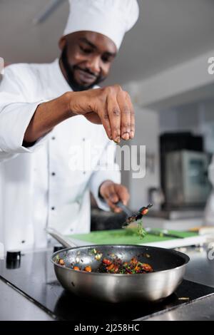 Chef cuisinier lancer des herbes fraîches hachées dans la casserole pour améliorer le goût du repas pendant que dans la cuisine professionnelle. Plat d'assaisonnement du chef principal préparé pour un concours de nourriture organisé dans un restaurant gastronomique. Banque D'Images