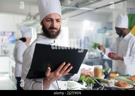 Travailleur de l'industrie alimentaire ayant un ordinateur portable, suivant une recette de plat gastronomique sur écran en remuant les ingrédients dans la casserole. Chef cuisinier avec ordinateur préparant la garniture pour le plat principal pendant que dans la cuisine du restaurant. Banque D'Images