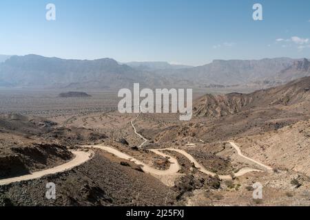 Une route de terre tortueuse qui mène en bas d'une montagne à l'étendue plate du Ghubrah Bowl, entouré par les montagnes arides de l'est de Hajar en Oman Banque D'Images