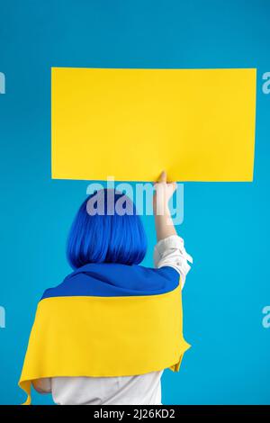 Soutenir le concept de l'Ukraine. Femme avec perruque bleue à cheveux courts, blouse blanche et drapeau national de l'Ukraine sur ses épaules debout avec affiche blanche jaune dans Banque D'Images