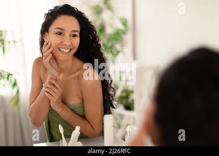 Charmante femme millénaire dans une serviette touchant son visage avec une peau rayonnante près du miroir dans la salle de bains, espace vide Banque D'Images