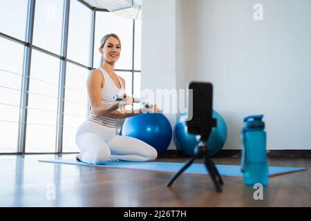 Fitness fille faisant des exercices avec des haltères assis sur le tapis et regardant dans le smartphone Banque D'Images