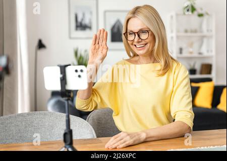 Femme sénior aux cheveux gris tenant un téléphone portable et se portant la main. Heureuse femme mûre souriante assise dans la cuisine, relaxante et parlant à son ami ou à sa famille via la connexion vidéo Banque D'Images