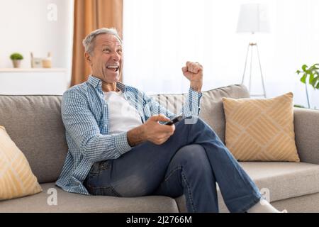 Un homme senior enthousiaste regardant la télévision célébrant la télécommande de maintien de l'objectif Banque D'Images