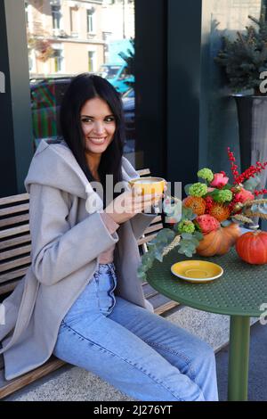 Belle jeune femme ukrainienne dans des vêtements décontractés d'automne buvant du cappuccino et souriant dans un café. Banque D'Images