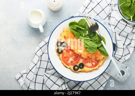 Omelette aux tomates, olives noires, fromage cottage et herbes aux épinards verts sur plaque blanche sur fond de béton gris. Délicieux petit déjeuner. En bonne santé Banque D'Images