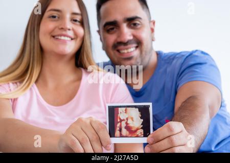 femme et homme caucasien enceinte souriant regardant l'appareil photo brouillé dans l'arrière-plan montrant la photo d'échographie imprimée, jeune couple hétérosexuel h Banque D'Images