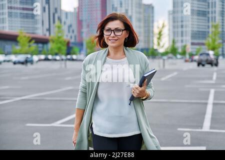 Souriante femme d'affaires mûre et mûre marchant à l'extérieur, arrière-plan moderne de style urbain. Banque D'Images
