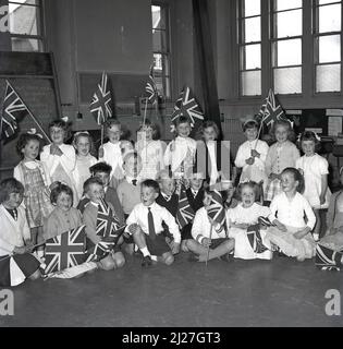 1965, historique, groupe d'enfants enthousiastes de l'école primaire assis dans une salle d'école tenant des drapeaux de Union Jack, Fife, Écosse, Royaume-Uni. Banque D'Images