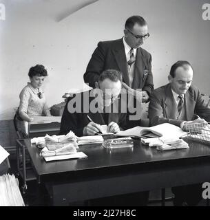 1950s, historique, travail de bureau, un directeur d'entreprise qui supervise le travail de deux commis de comptes hommes matures ou des comptables assis les uns à côté des autres à un bureau, Abbey Works, Port Talbot, pays de Galles, Royaume-Uni. Également au bureau, une femme secrétaire avec machine à écrire. Banque D'Images