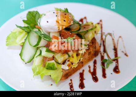 Griller avec du saumon et de l'œuf poché. Laitue, concombre, basilic. Un sandwich santé pour le petit déjeuner. Graines de Microgreen et de lin et de sésame. Aliments sains sur une assiette blanche. Poisson avec légumes sur le pain. Banque D'Images