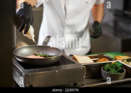 Vue en crêpe des mains du chef masculin, verser de l'huile d'olive sur du cutlet juteux avec du romarin dans une casserole. Préparation appétissante, hachée au grill pour hamburger de bœuf avec des ingrédients sur fond. Concept de cuisine. Banque D'Images