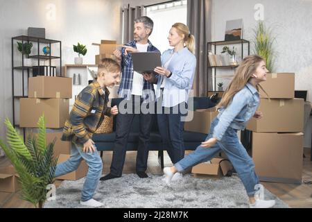 Deux enfants amusants, garçons et filles, jouer, courir et s'amuser pendant la réinstallation, tandis que les parents se sont concentrés derrière l'utilisation d'un ordinateur portable et discuter de la conception dans un appartement moderne. Banque D'Images