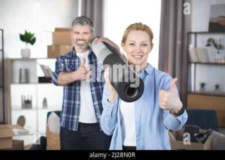 Un couple caucasien souriant portant un tapis roulé après s'être déplacé dans une maison. Concentrez-vous sur une jolie femme blonde souriante, tenant le tapis sur son épaule et montrant le pouce vers le haut et son agréable homme barbu derrière Banque D'Images