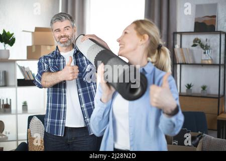 Un couple caucasien souriant portant un tapis roulé après s'être déplacé dans une maison. Concentrez-vous sur l'homme barbu agréable derrière regarder sur sa femme jolie femme blonde, montrant le pouce et souriant, tenant le tapis de rouleau Banque D'Images