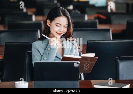 Femme entrepreneur d'Asie montrant un visage souriant en lisant un livre développant des stratégies financières et d'investissement et en utilisant une tablette d'ordinateur Banque D'Images