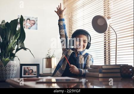 L'apprentissage ne s'arrête jamais, même en verrouillage. Photo d'un adorable petit garçon utilisant un casque et levant la main tout en terminant une mission scolaire Banque D'Images