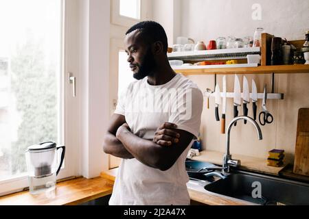 Homme attentionné avec bras croisés regardant vers le bas tout en s'appuyant sur le comptoir de cuisine à la maison Banque D'Images