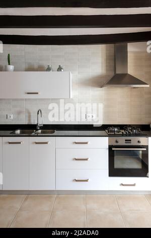 Cuisine blanche avec lumière naturelle. Mobilier blanc en marbre noir. Plafond avec poutres en bois. Table de cuisson à gaz et ventilateur d'extraction en acier inoxydable. Banque D'Images