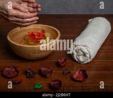 Les mains des femmes avec de l'eau s'écoulant dans un bol en bois avec une fleur jaune et rouge à l'intérieur, sur une table en bois avec une serviette enroulée Banque D'Images