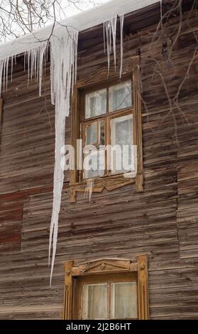 Des glaçons glacés transparents sont suspendus sur le bord du toit. Sur le fond du mur en bois de la vieille maison. Grandes cascades, même de belles rangées. Jour d'hiver nuageux, lumière douce. Banque D'Images