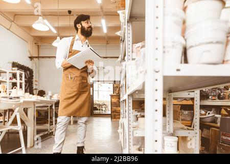 Jeune homme d'affaires séduisant un potter avec une barbe et une moustache travaille dans son atelier. Conserve les enregistrements et transcrites dans un ordinateur portable en inspectant Banque D'Images