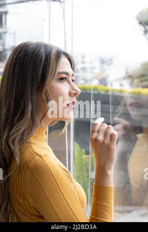 belle femme latine avec de longs cheveux blonds regardant par la fenêtre à la ville, elle porte le maquillage et les vêtements colorés, la beauté et le style de vie modèle Banque D'Images